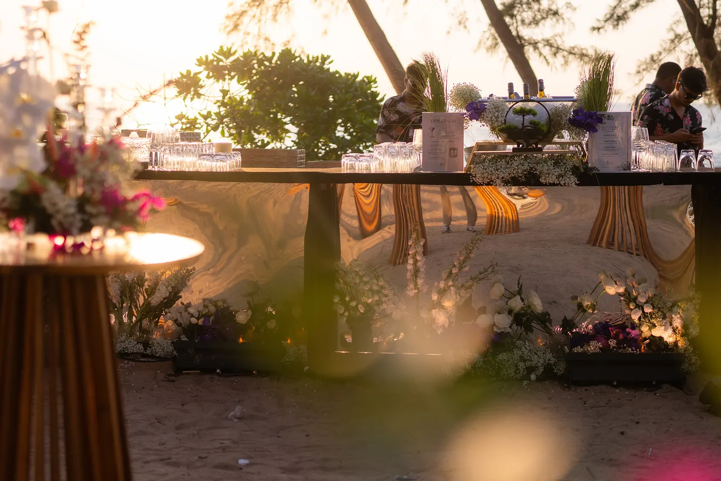 Wedding Setup — mirrored beach bar station dressed with glassware and purple and white flowers at sunset — ภาพงานแต่งงานและคู่บ่าวสาว