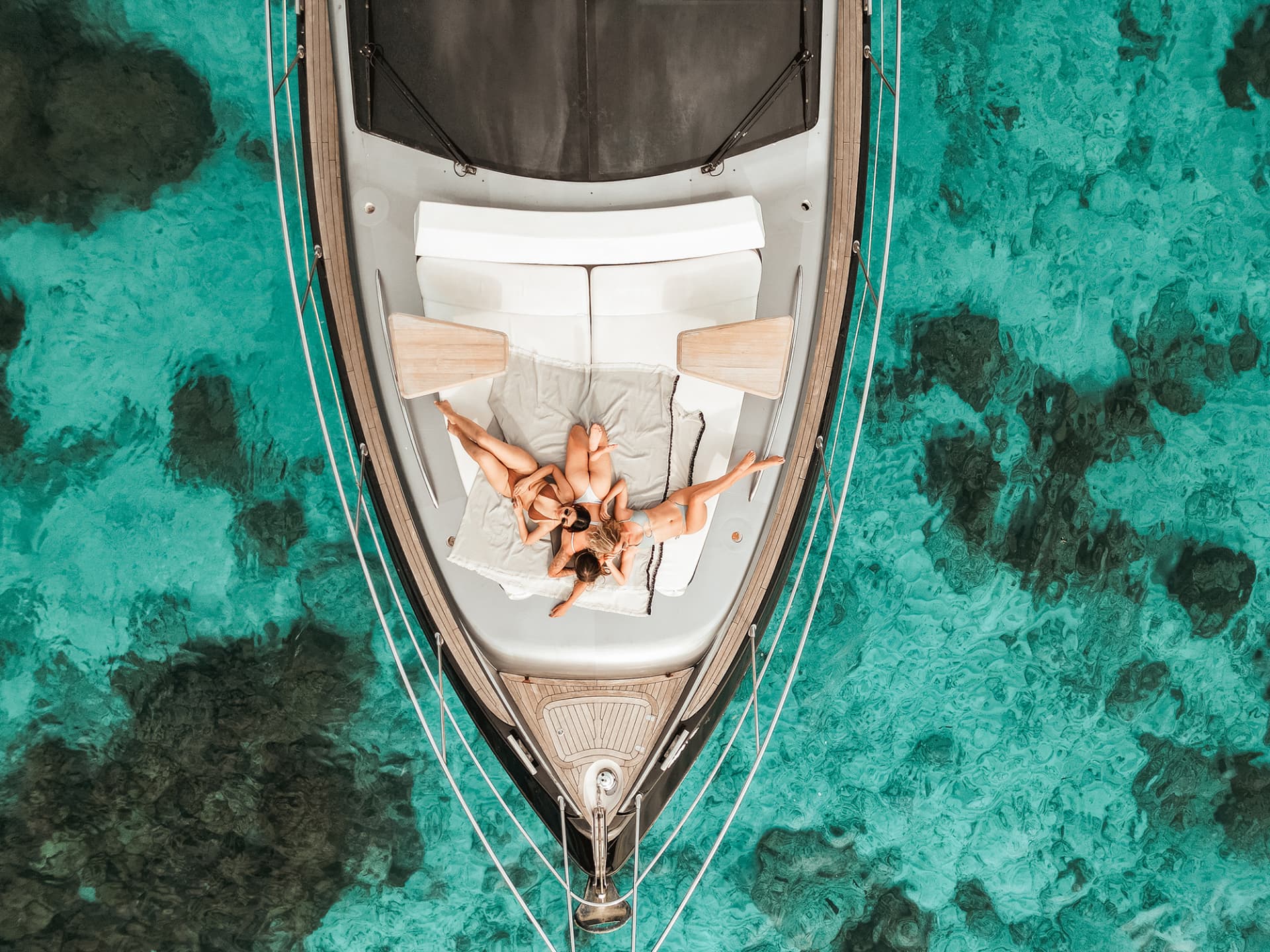 Hype Yacht — top-down aerial of three guests on the bow sunpad above a coral reef