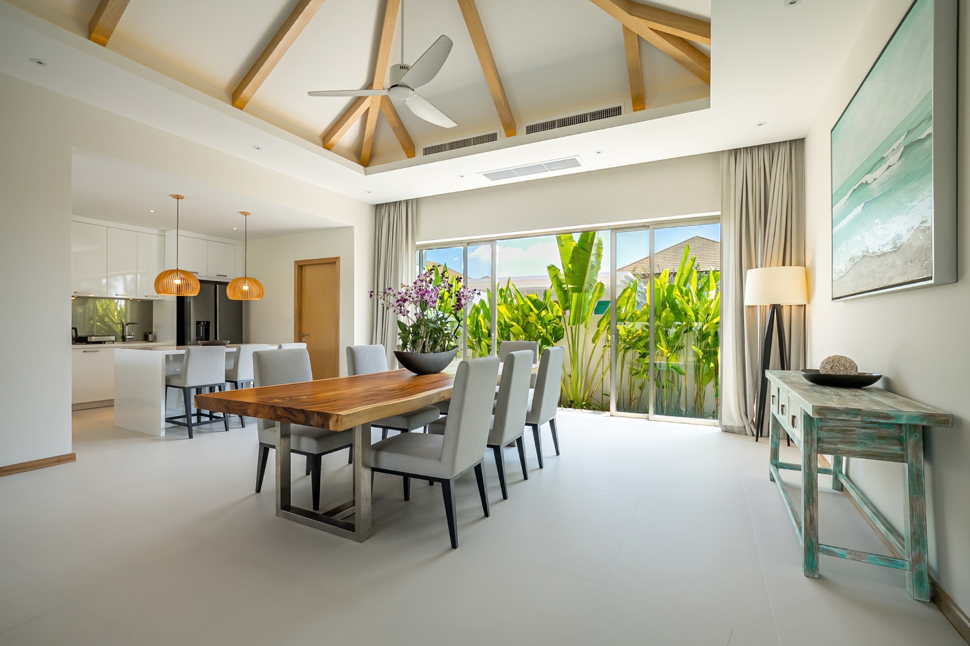Trichada Villas — dining room with a live-edge timber table under a vaulted beam ceiling, opening to the kitchen and garden