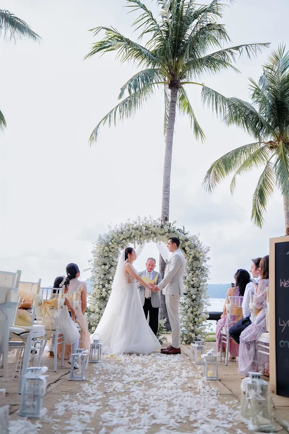 Thomas & Jane The Wedding — ceremony under a white floral arch between palm trees overlooking the sea — ภาพงานแต่งงานและคู่บ่าวสาว