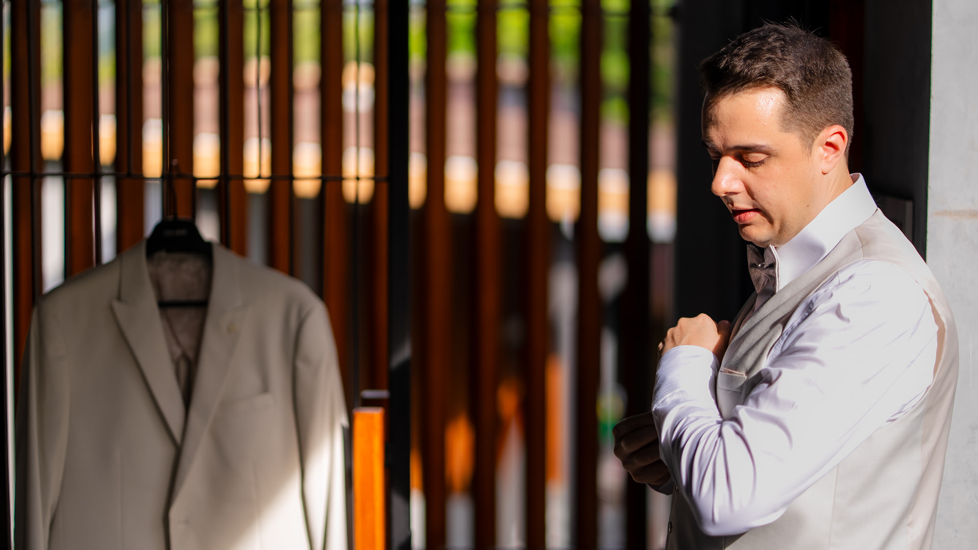 Thomas & Jane The Wedding — groom fastening his boutonnière beside a hanging jacket in warm evening light
