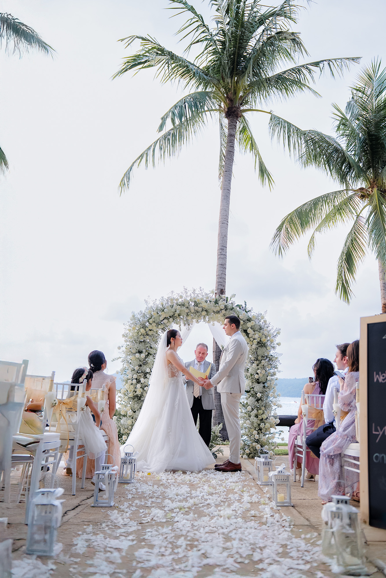Thomas & Jane The Wedding — ceremony under a white floral arch between palm trees overlooking the sea