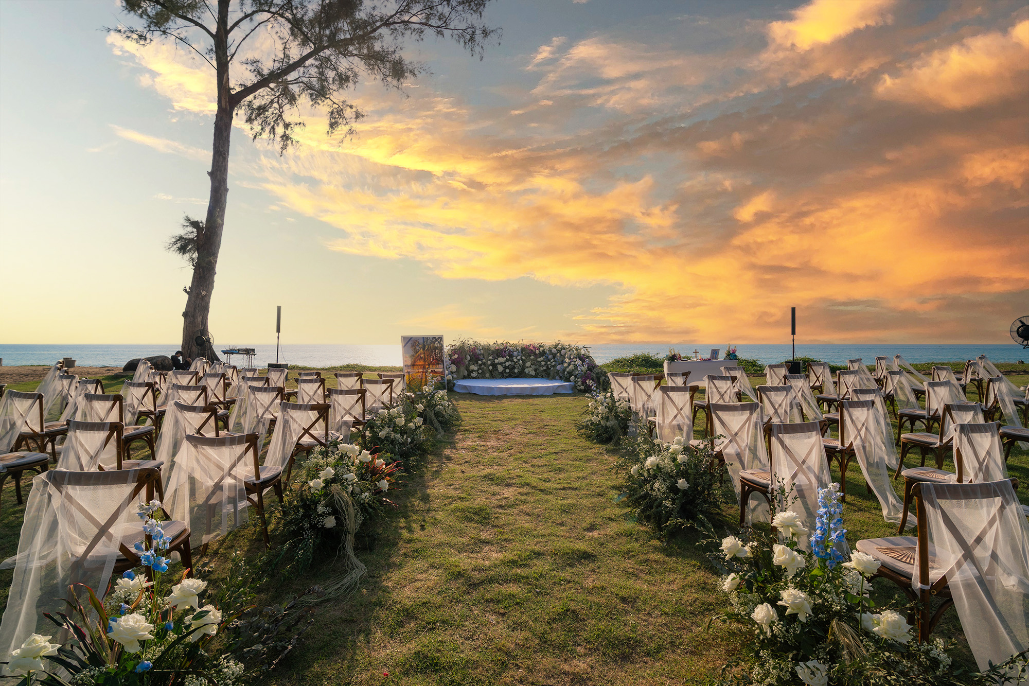 Wedding Setup — seaside ceremony aisle on the lawn leading to a circular white altar and floral arch under a sunset sky
