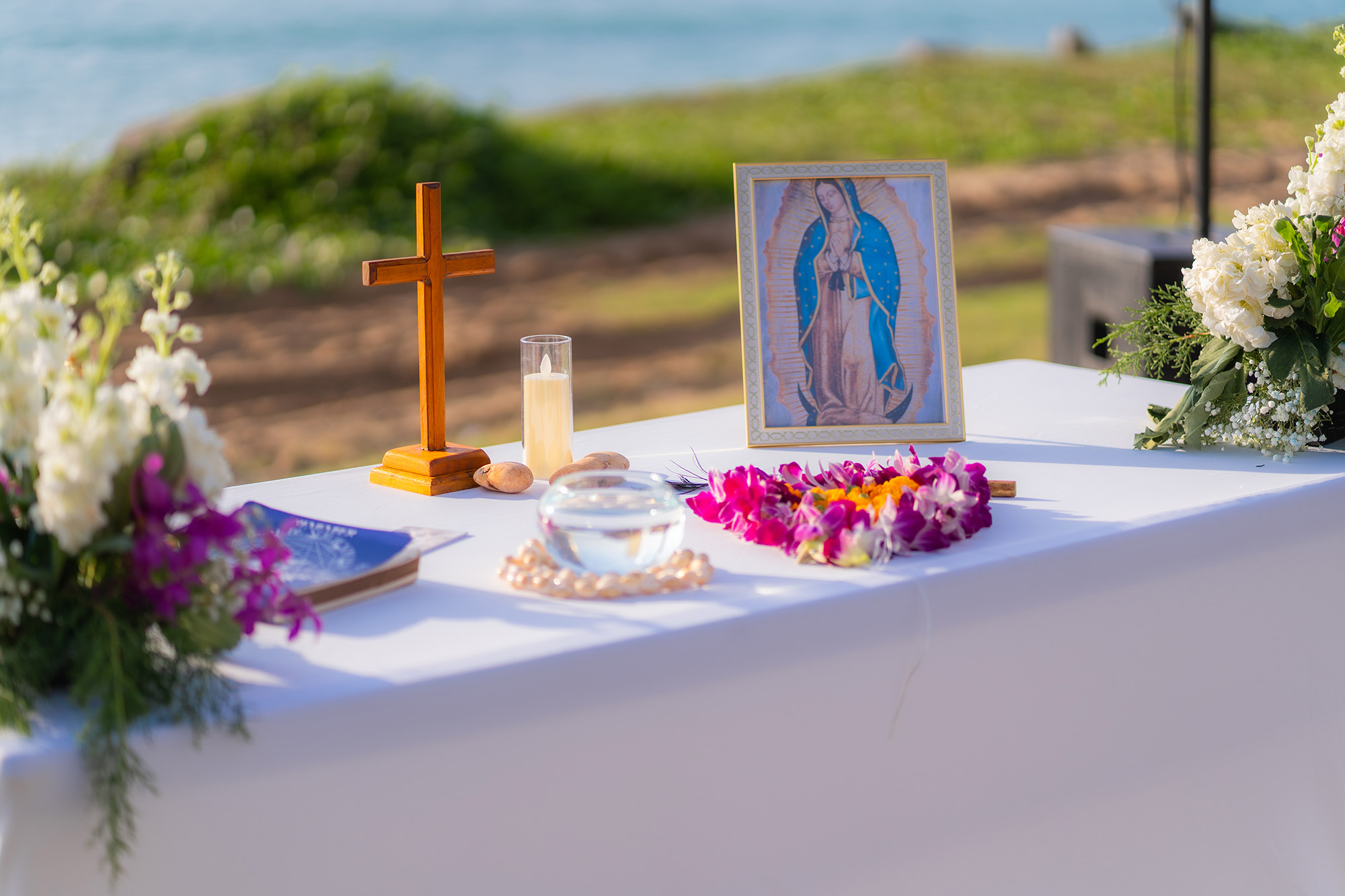 Wedding Setup — altar table before the sea with a wooden standing cross, a cased candle and a framed portrait of Our Lady of Guadalupe