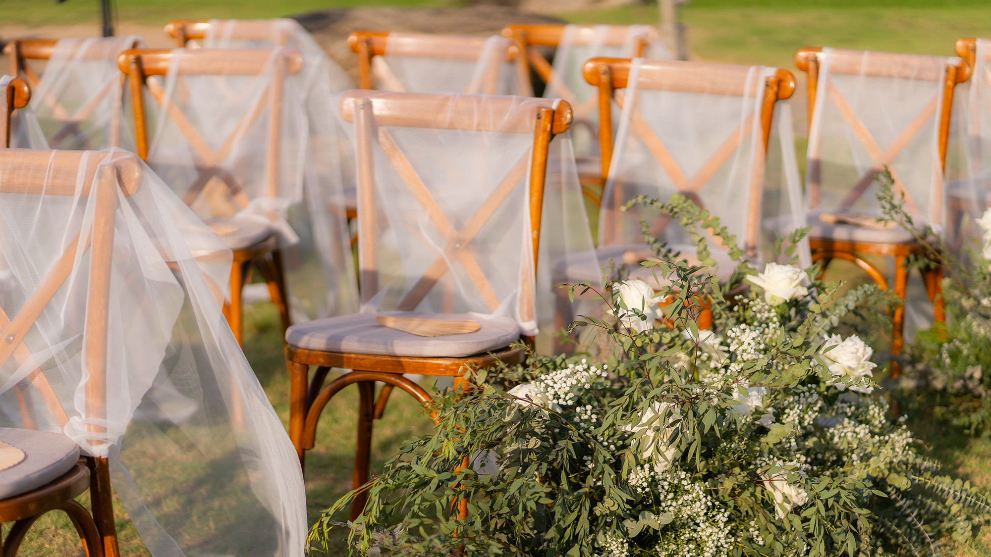 Wedding Setup — lawn ceremony aisle of wooden chairs draped in sheer white tulle beside an arrangement of eucalyptus, white roses and baby's breath